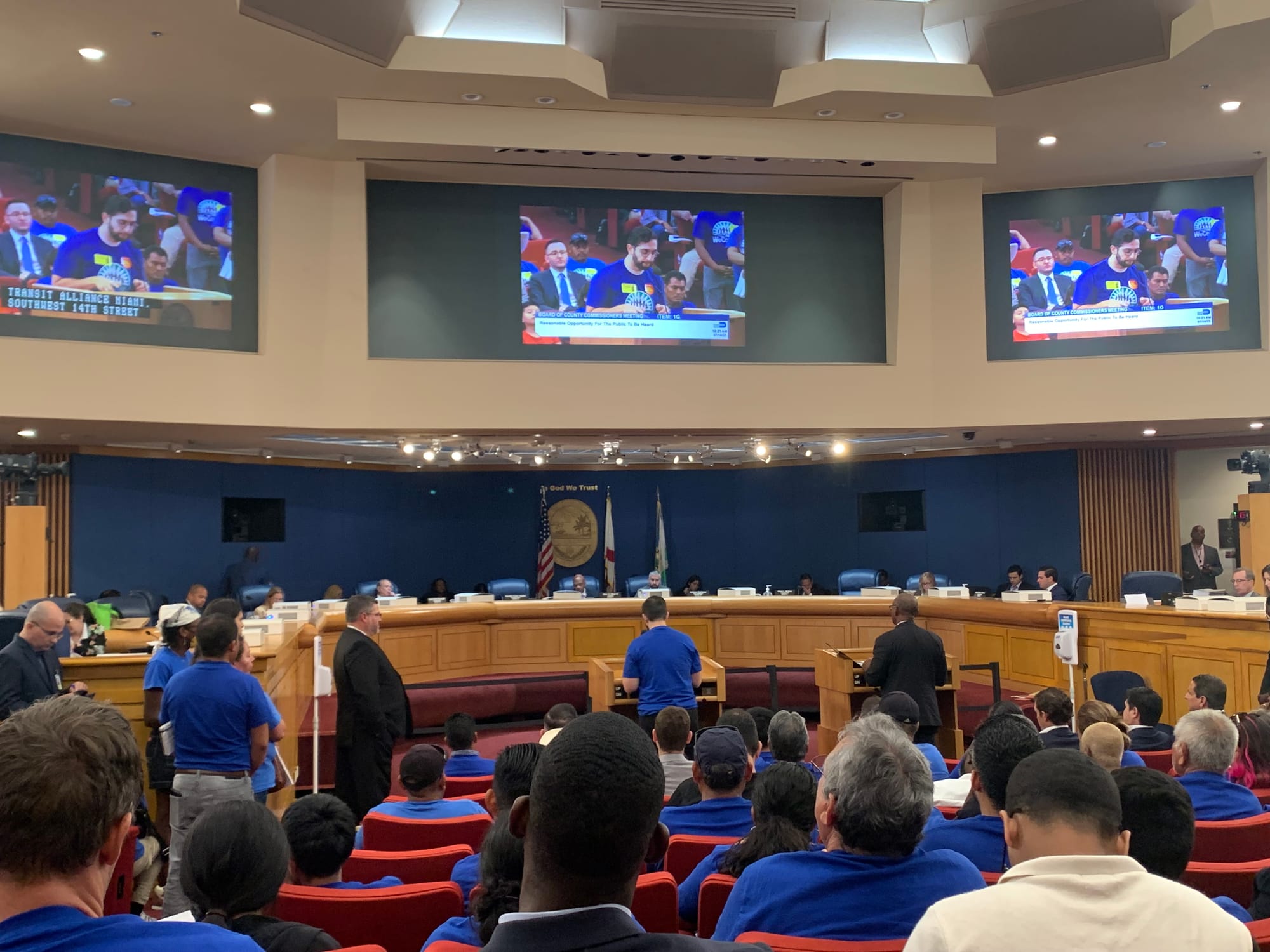 color photograph inside a public government building. people in blue shirts sit in auditorium seats in front of a panel of commissioners. three large screens above the commissioners' heads show a person in a blue shirt speaking at a podium