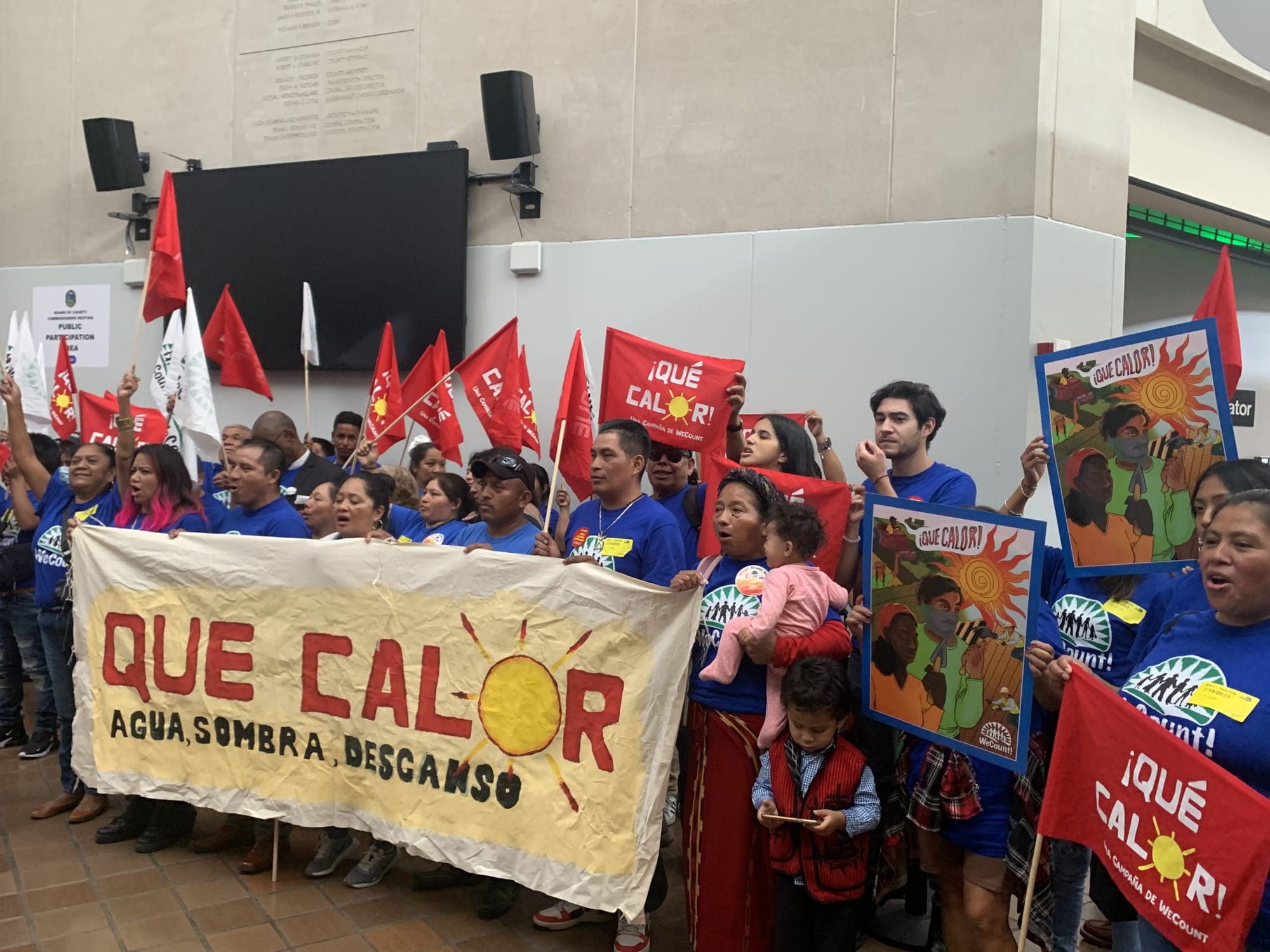 color photograph of an outdoor protest. people hold red cloth signs with white texts that reads "¡Que Calor!" and several peo