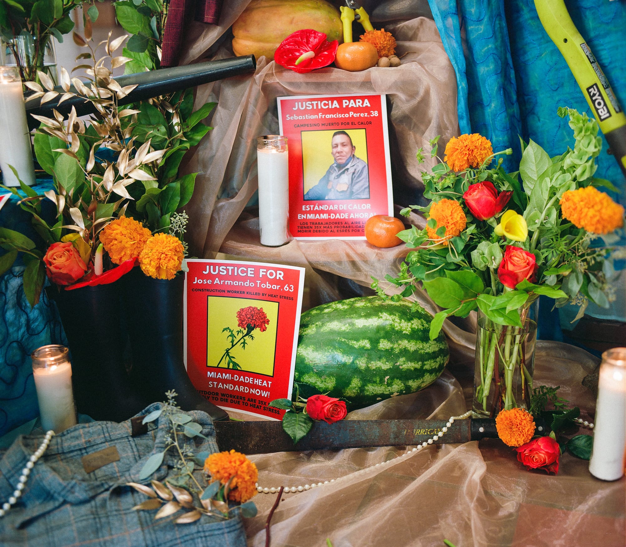 color photograph of an outdoor altar cover in light brown sheer cloth. flowers in hues of red, orange, and yellow decorate the altar, along with watermelon and posters printed with bright red backgrounds and white text that calls for justice for farmworkers who have died