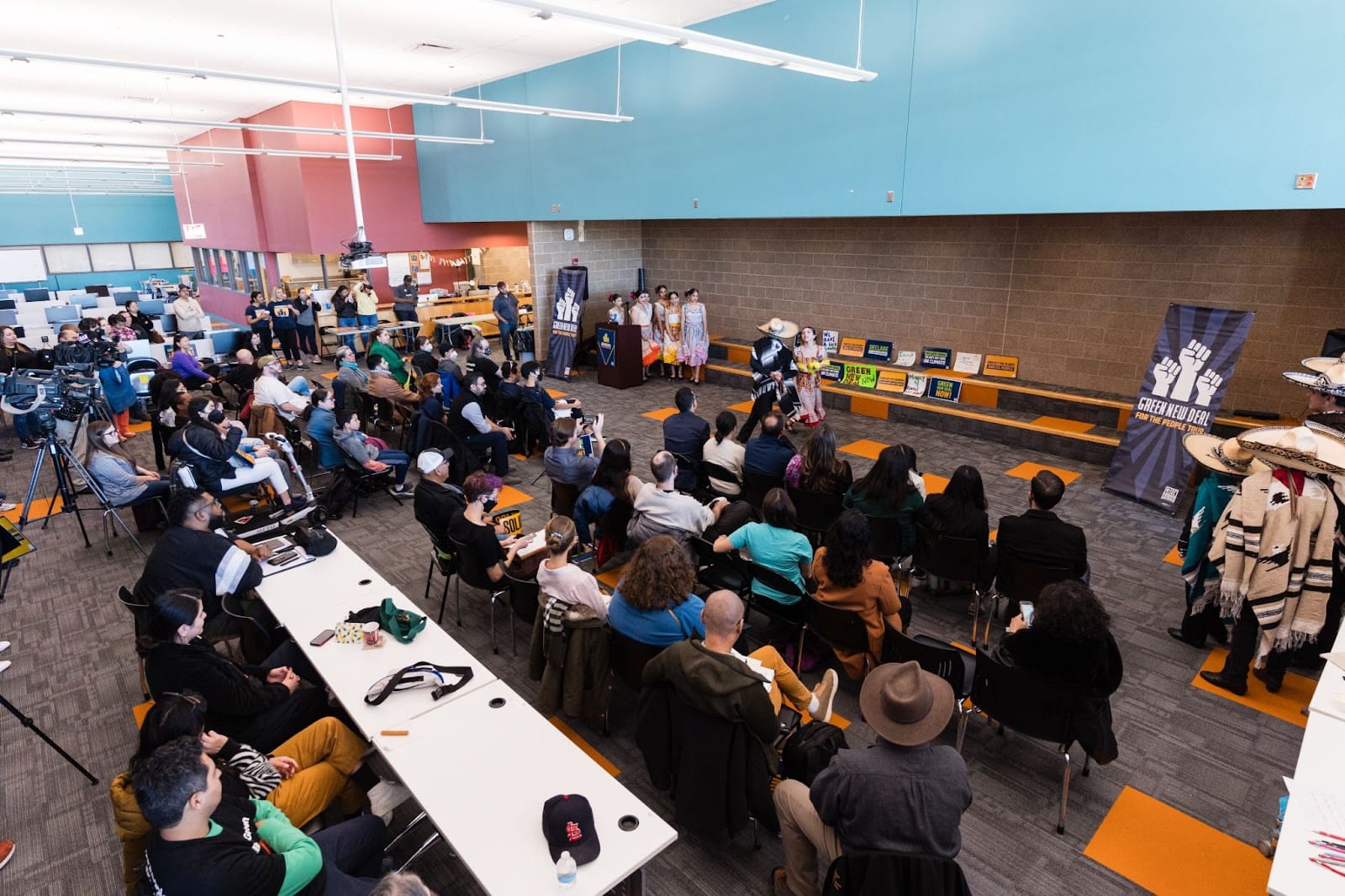 color photograph of people sitting at long tables and rows of chairs facing the front of a school media center where a skit is being performed. in the background, there is a vertical banner with three fists raised in solidarity and text that reads "green new deal for the people tour"