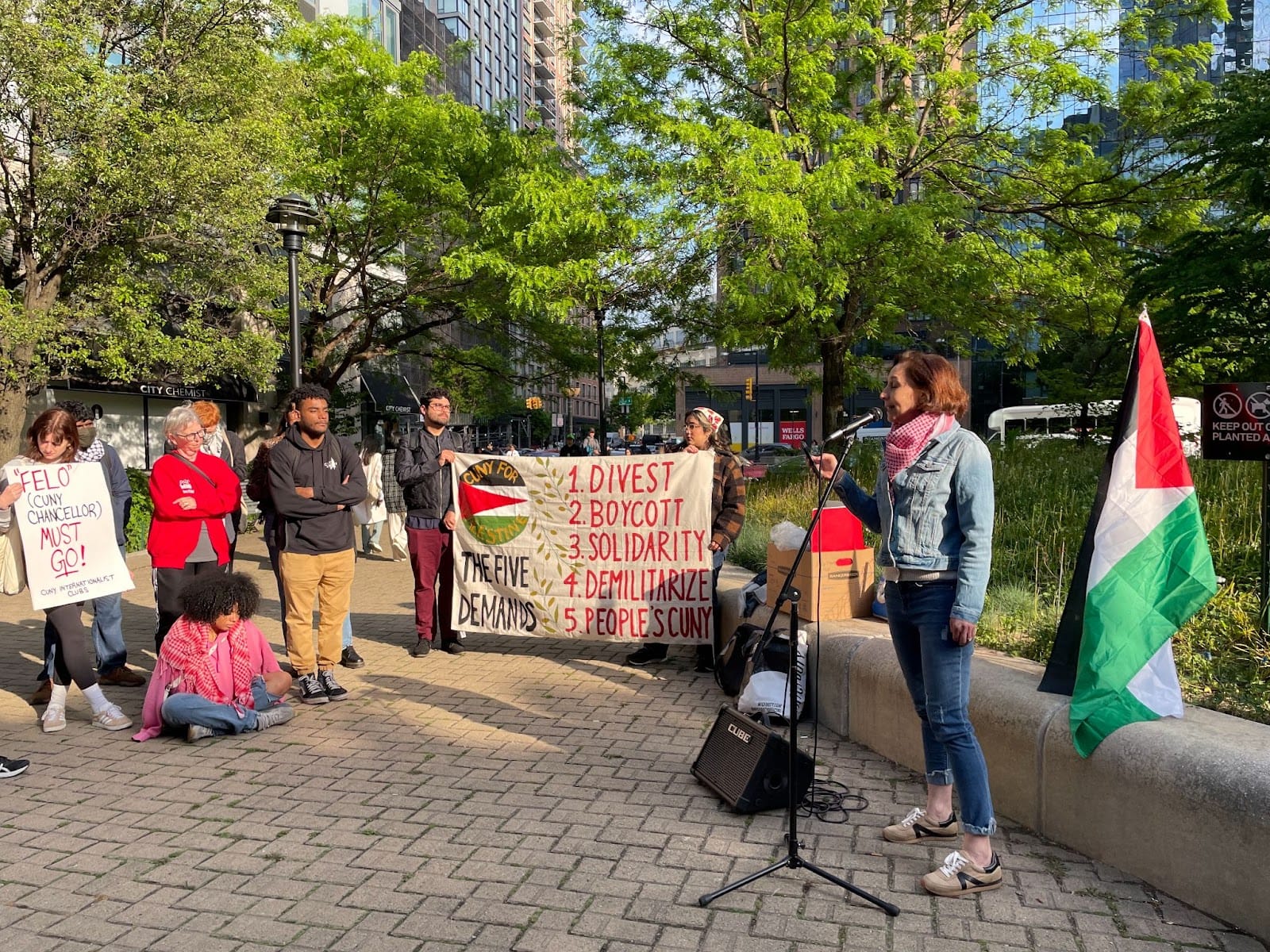 color photograph of a woman wearing a red and white keffiyah speaking at a microphone on a stone sidewalk. two people hold a banner reading "1. divest 2. boycott 3. solidarity 4. militarize 5. people's cuny"