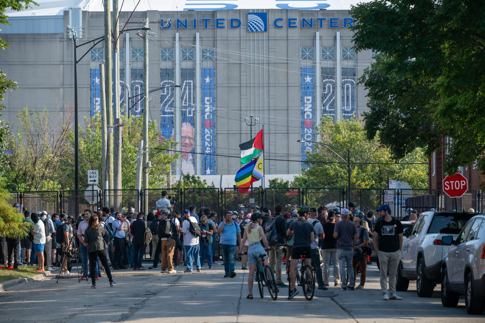 color photograph of people holding up Palestinian and Pride flags in front of fencing in the mid-ground. in the background is the United Center with banners of Tim Walz's face and DNC 2024 logos