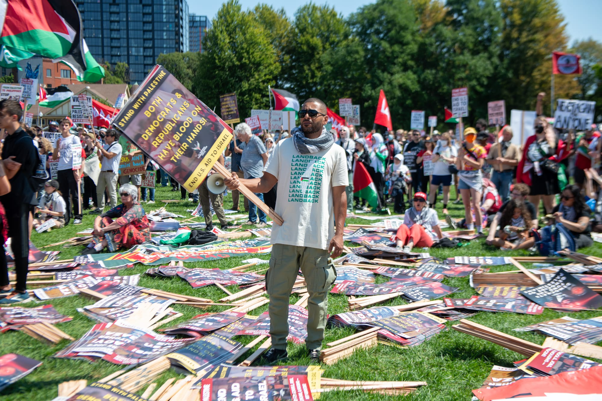 color photograph of people resting picket signs on grass. a man wearing a LANDBACK shirt and keffiyeh stands in the center with a picket sign that reads "both Democrats and Republicans have blood on their hands"