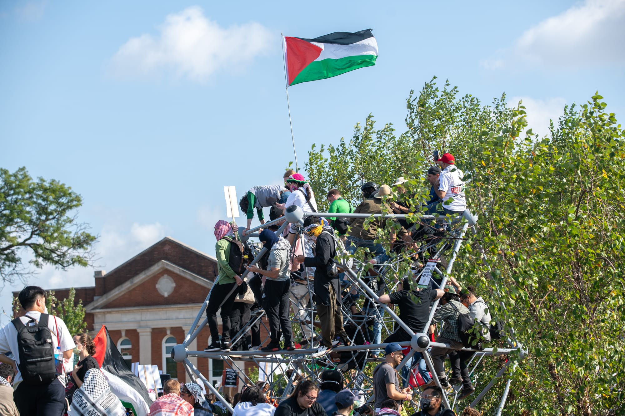 color photograph of people climbing a jungle gym structure and waving a Palestinian flag from the top