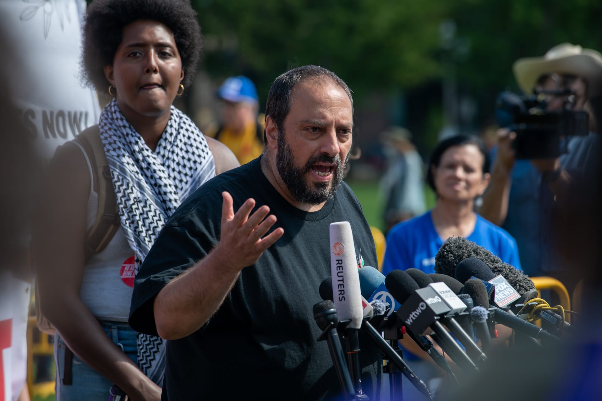 color photograph of a Middle Eastern man standing at a podium and gesturing as he speaks. Behind him are several people watching him as he speaks