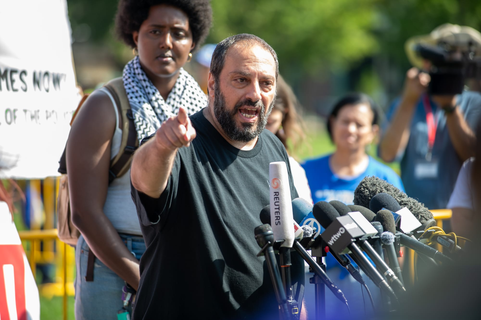 color photograph of a Middle Eastern man standing at a podium and pointing toward the camera as he speaks. Behind him are several people watching him as he speaks
