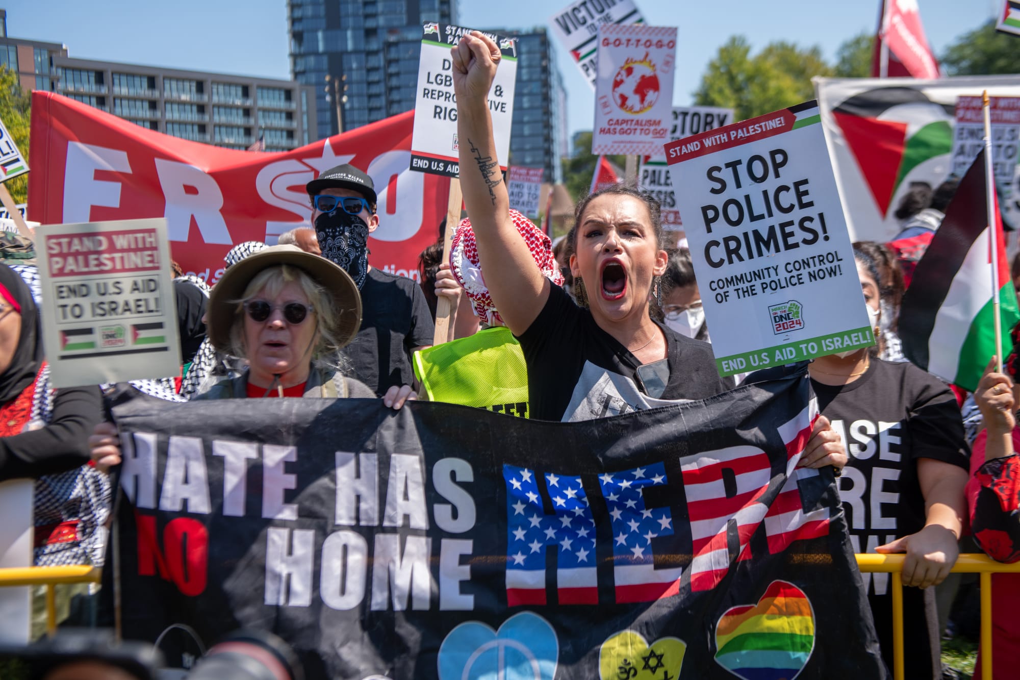 color photograph of people holding up signs and banners. in the foreground, a femme person holds up their fist as they hold a black banner that reads "hate has no home here"