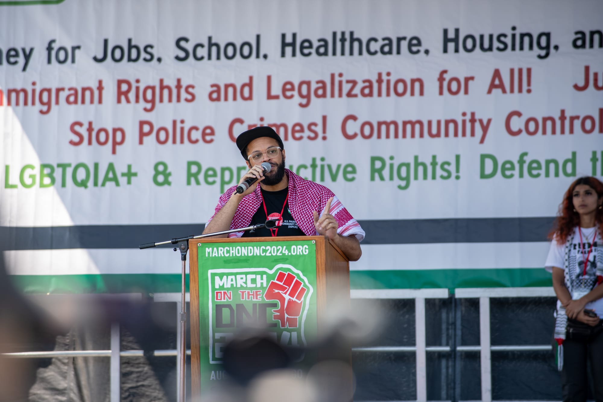 color photograph of a man wearing a red and white keffiyeh speaking at a podium on stage in front of a large banner with pro-labor, pro-health care, and pro-education slogans
