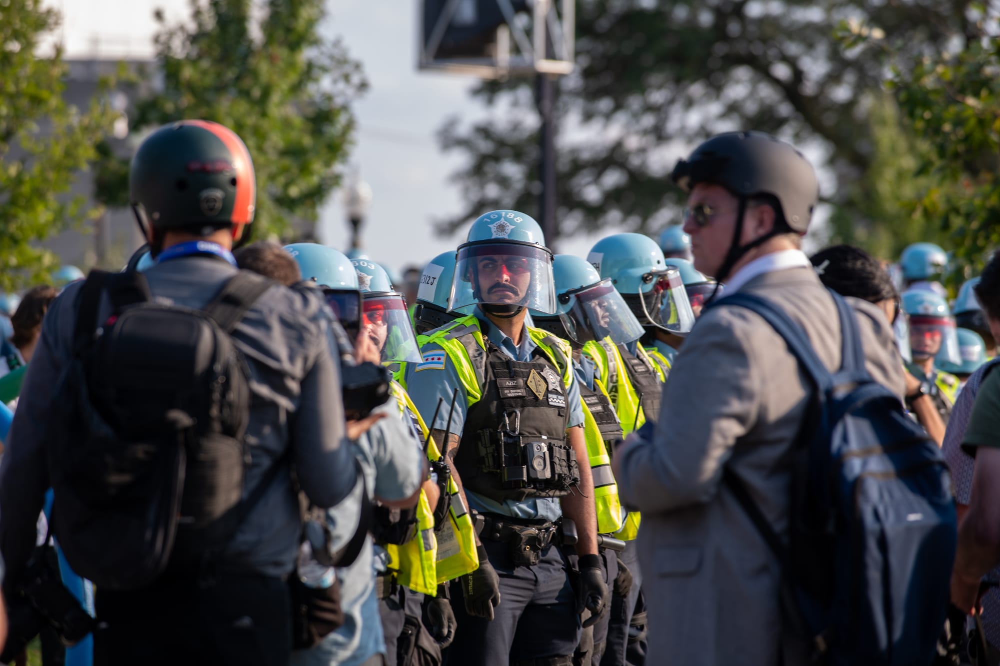 color photograph of protesters in suits and bicycle helmets in the foreground standing in front of a line of police officers