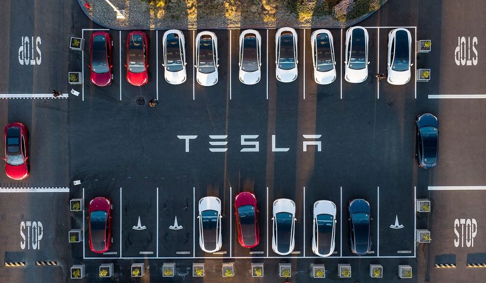 tesla vehicles are lined up in two parallel lines of parking spaces. between the two lines of parking spaces, the word "tesla