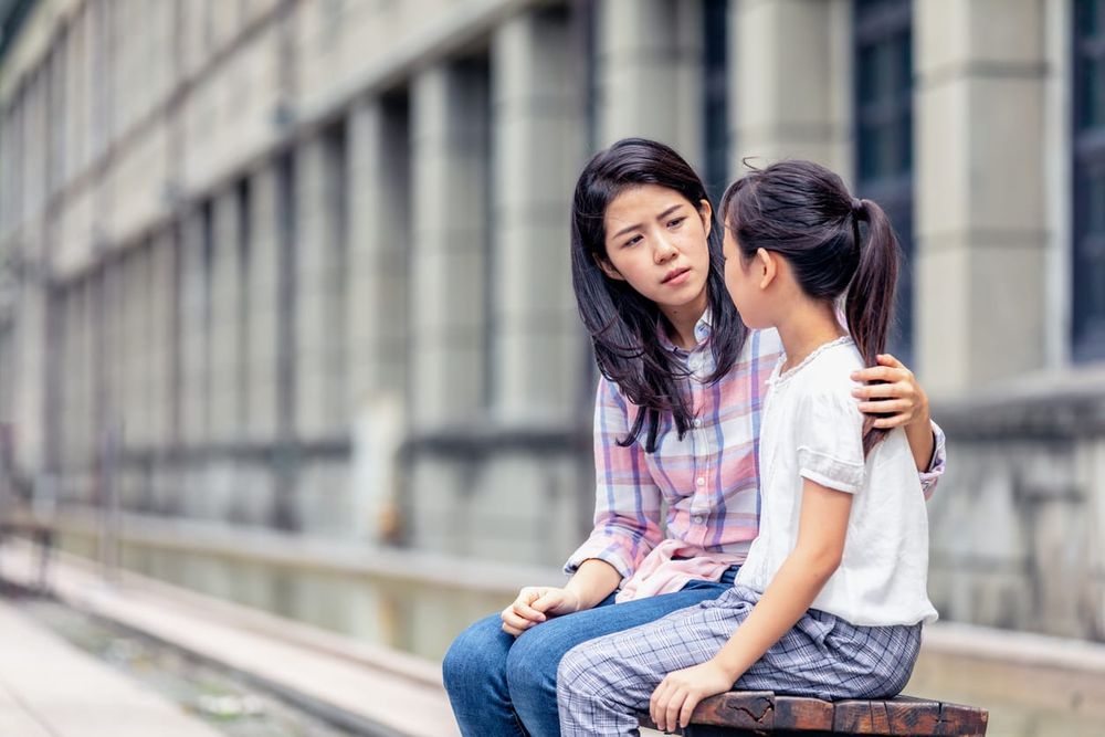 A young asian woman with dark hair sits with her arm around her child, looking serious. The child faces away from the camera.