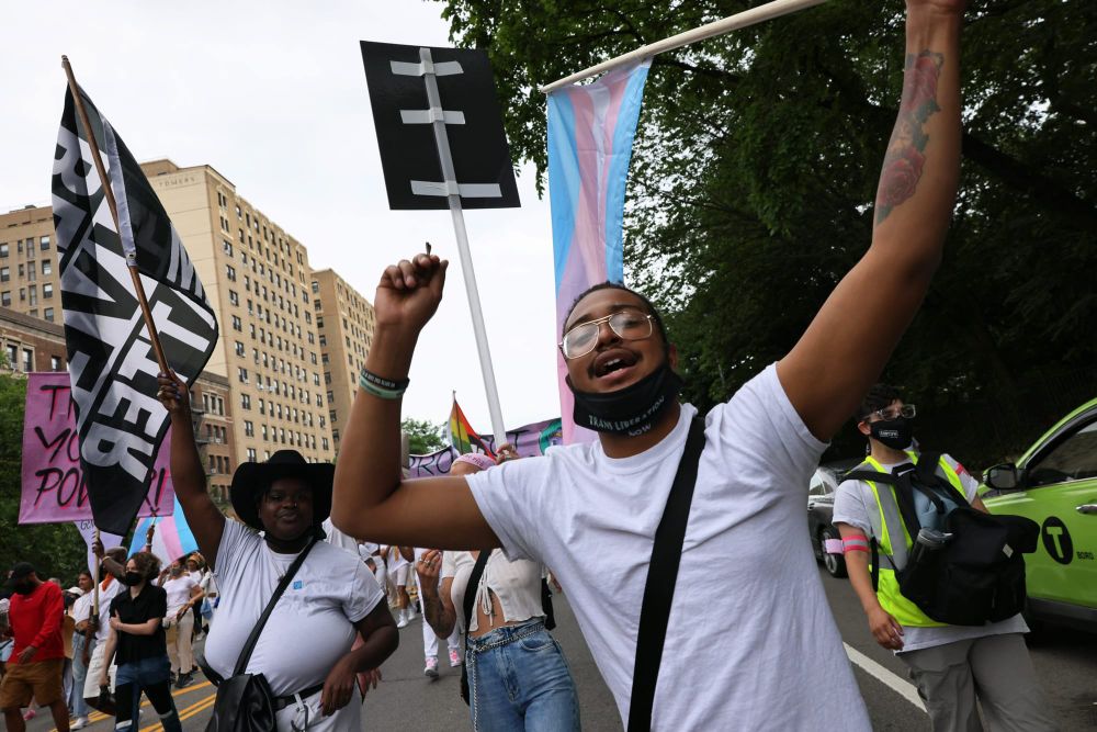 a Black person stands in the foreground and holds a trans flag as they walk down the street accompanied by other protesters