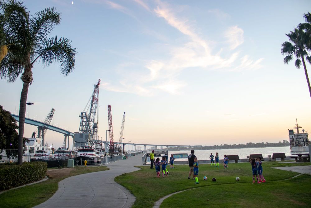 Latinx children play soccer with adults supervising on the field. In the background is a bay and cranes