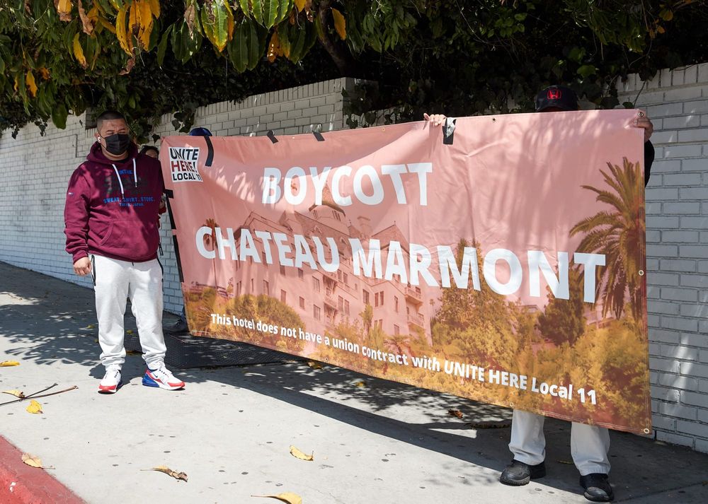 workers wearing red shirts and long white pants hold a peach-colored banner with serif white text reading "boycott chateau ma
