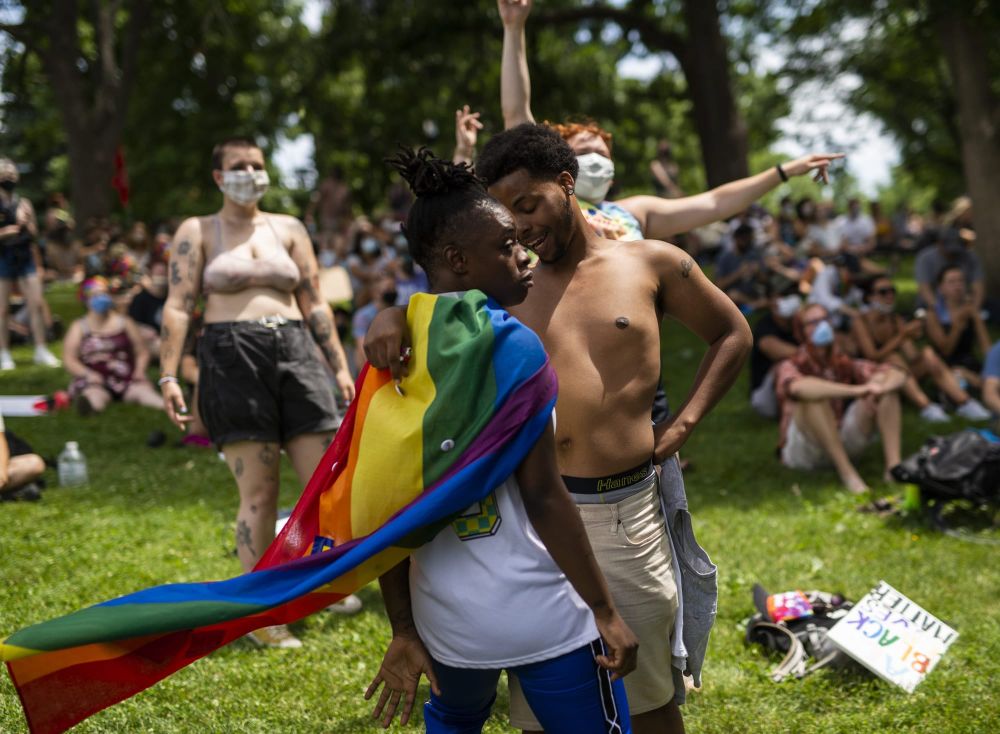 color photograph of a pride celebration in a grassy park with trees in the background. people sit and stand close together on