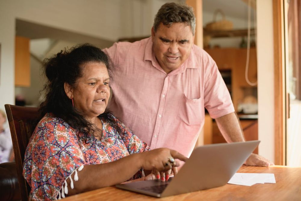color stock photo of an Aboriginal couple in their 50s at home using the internet. The man leans over woman and looks at the
