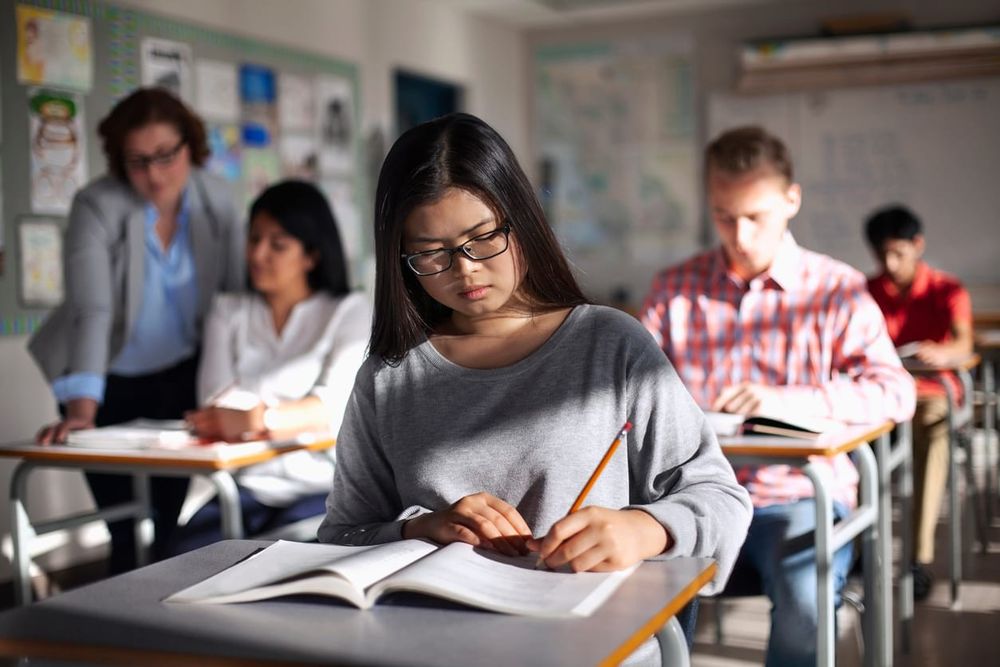 stock color photo of a Vietnamese girl in a long-sleeve grey sweater sitting at a classroom desk and writing in a notebook. b