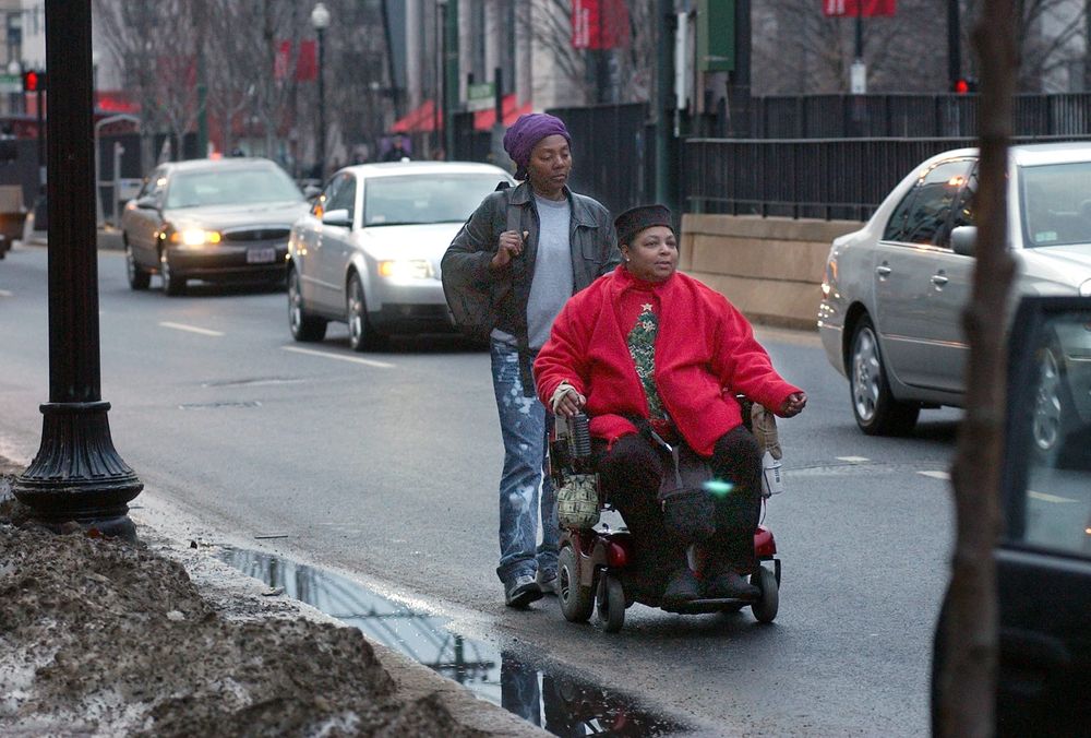 color photograph of a Black woman in a wheelchair traveling on a paved street. another Black woman walks behind her, and cars