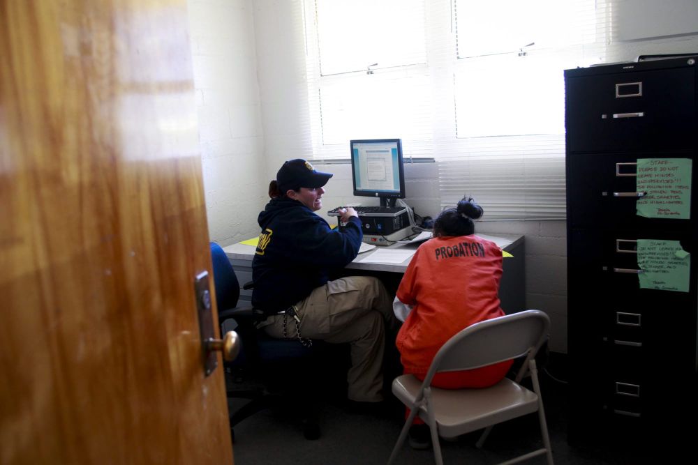 color photograph of a young girl in an orange jumpsuit with black text that reads "probation" across the shoulder blades. she
