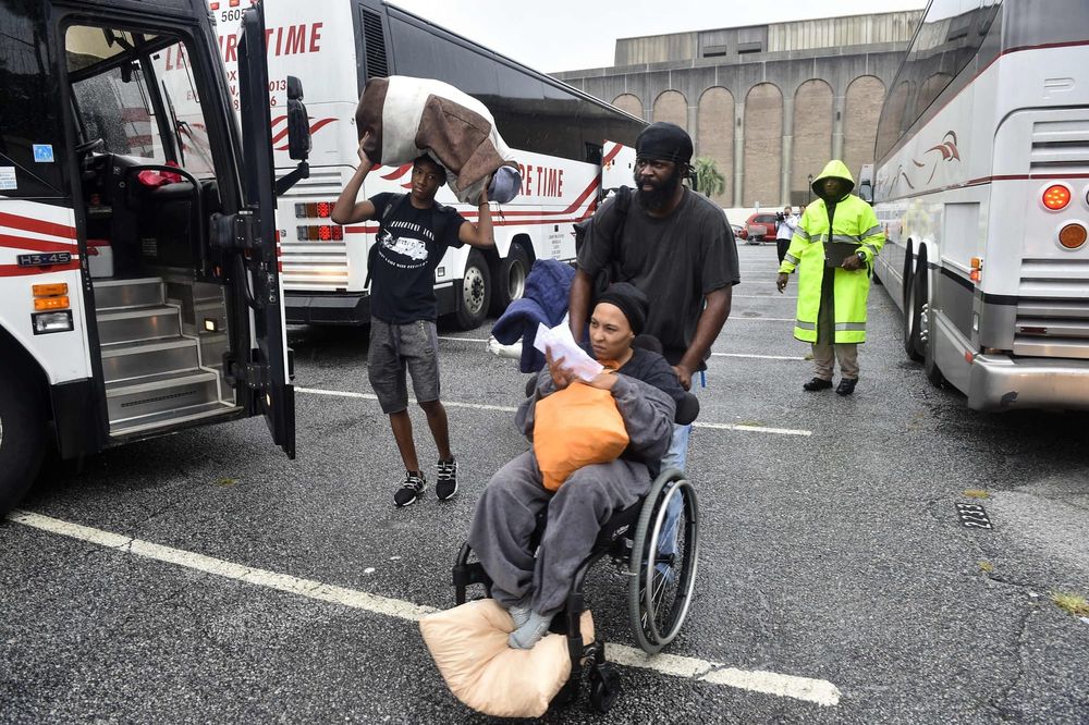 color photograph of a Black woman in a wheelchair being pushed by a Black man in a parking lot. several long buses surround t