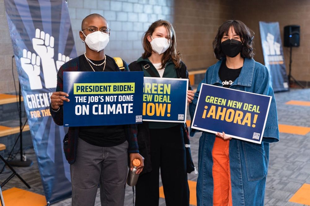 color photograph of three people standing inside wearing masks. they hold blue posters with printed text slogans supporting t