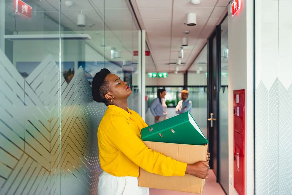 color photograph of a Black woman in a vibrant yellow shirt and white pants holding a cardboard box in the hallway of a corpo