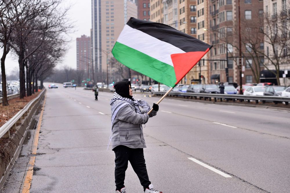 color photograph of a person wearing a black head covering waving a Palestinian flag in the middle of an empty street