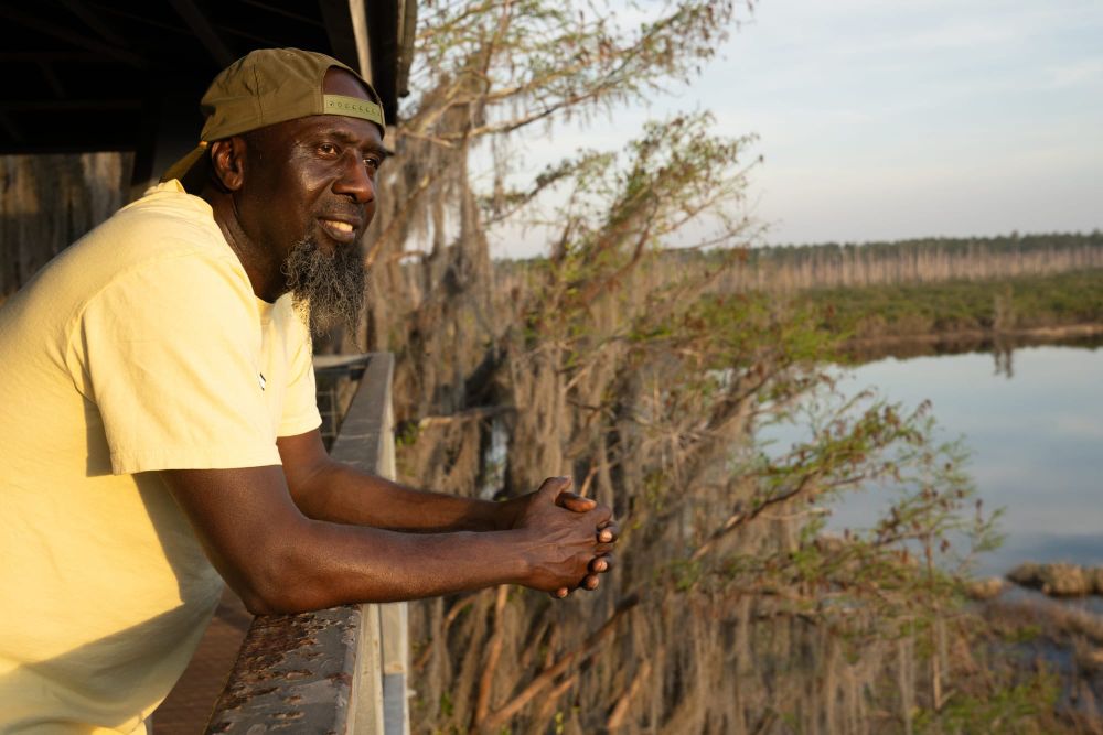 color photograph of a Black man in a light yellow t-shirt leaning on a wooden balcony overlooking a swamp
