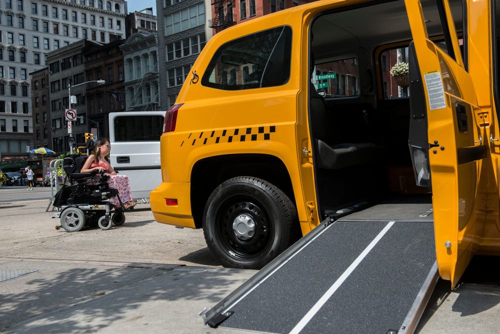 color photograph of a yellow taxi cab in new york city with the wheelchair ramp unfolded. a person in a wheelchair waits behi