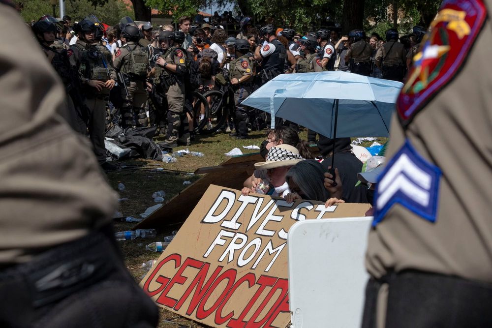 color photograph of two police officers' shoulders in the foreground framing a sign calling for divestment from genocide