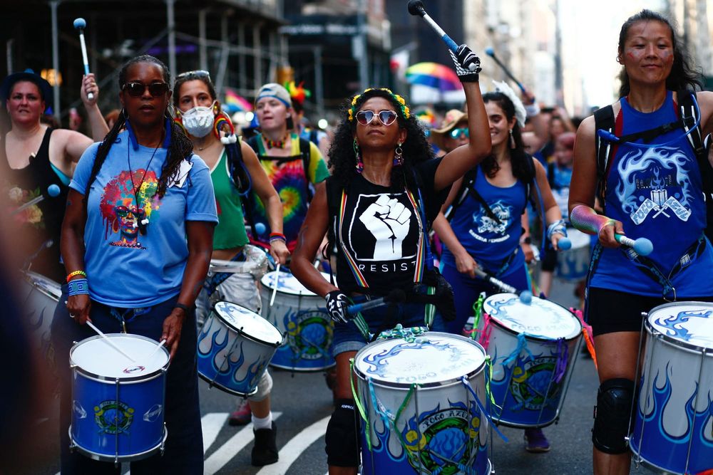 color photograph of an outdoor Pride celebration. Several Black people stand in the foreground in a drum line