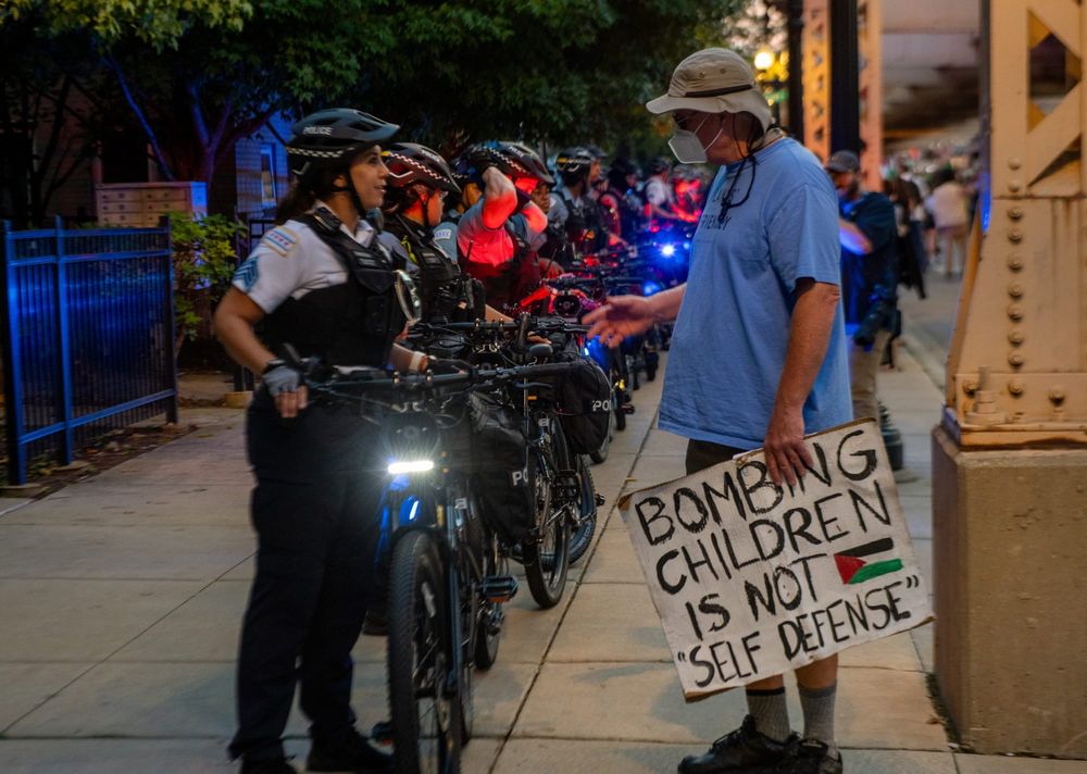 photo of a person in a blue shirt and tan bucket hat holding a handmade sign reading, "bombing children is not self defense"