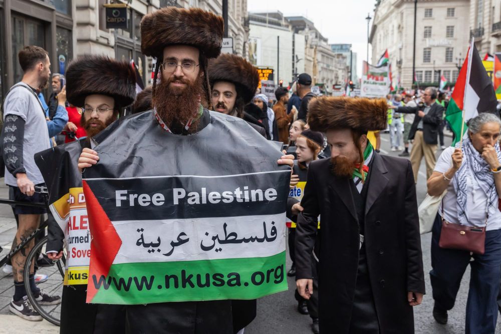 Protesters with beards and traditional Orthodox Jewish attire, one holding a sign that says "Free Palestine"