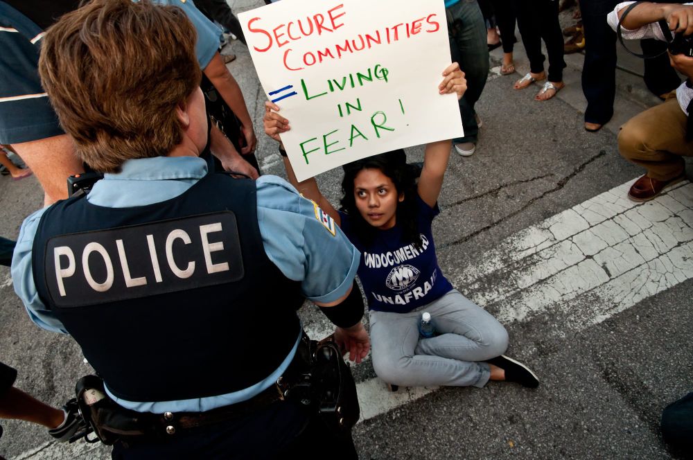 A woman sits on the street in front of a police officer; she's holding up a sign that says, "Secure Communities = Living in F