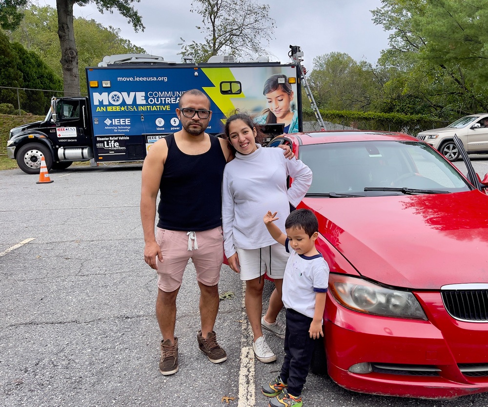 A man, woman and child smile at the camera as they stand next to a red car in a parking lot.