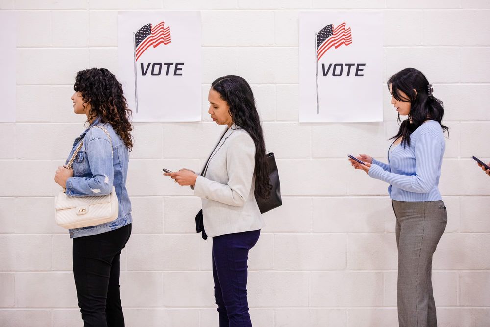 Young women look at their phones while standing in line to vote.