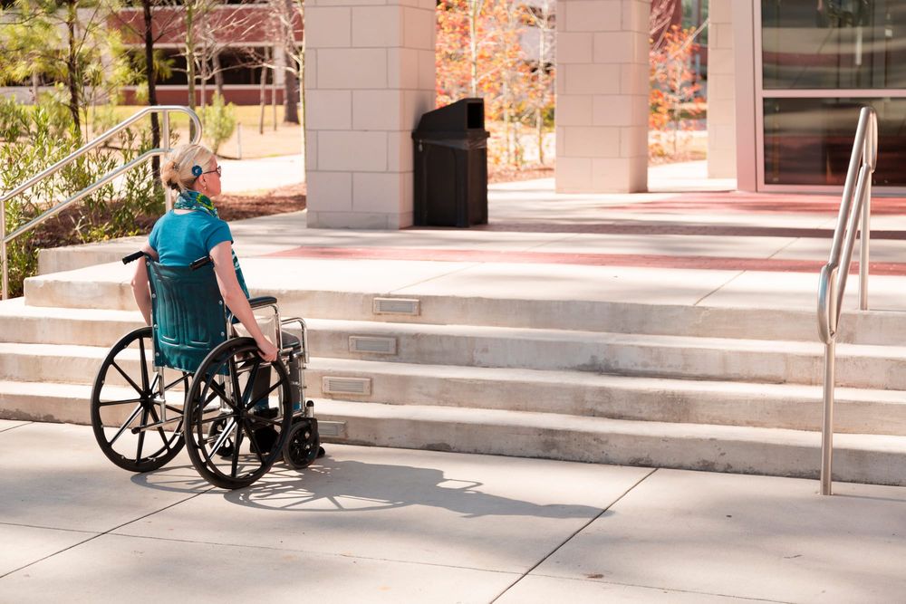 woman in wheelchair faces a flight of stairs leading into a building
