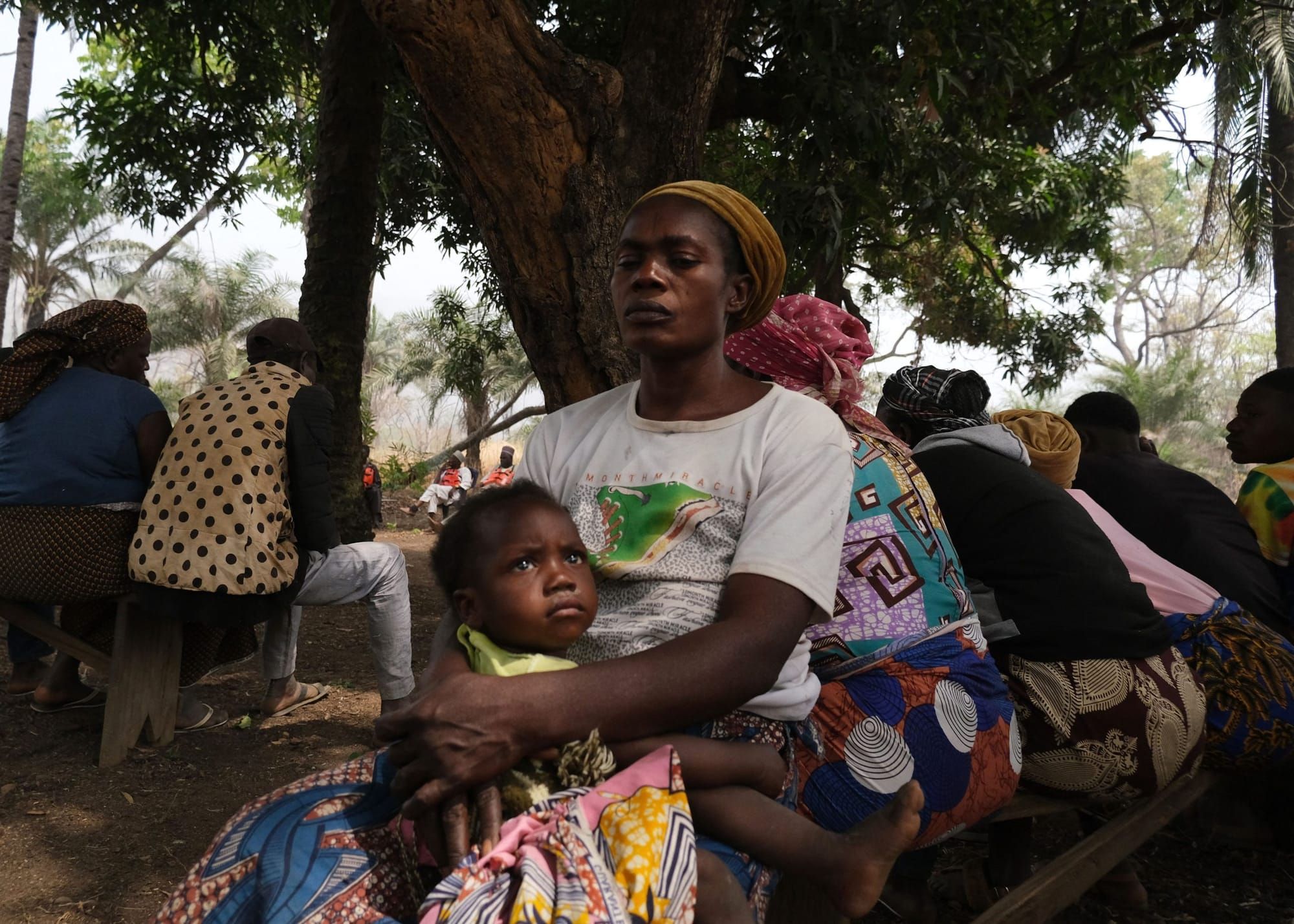 a Black woman wearing a head covering sits on a bench with a child in her lap facing the camera. Seven other people sit with