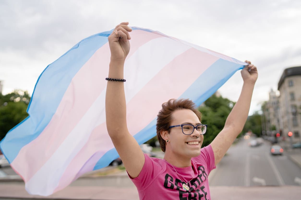 A person holding a transgender flag waving in the air is wearing a bright pink t-shirt outside on a cloudy day
