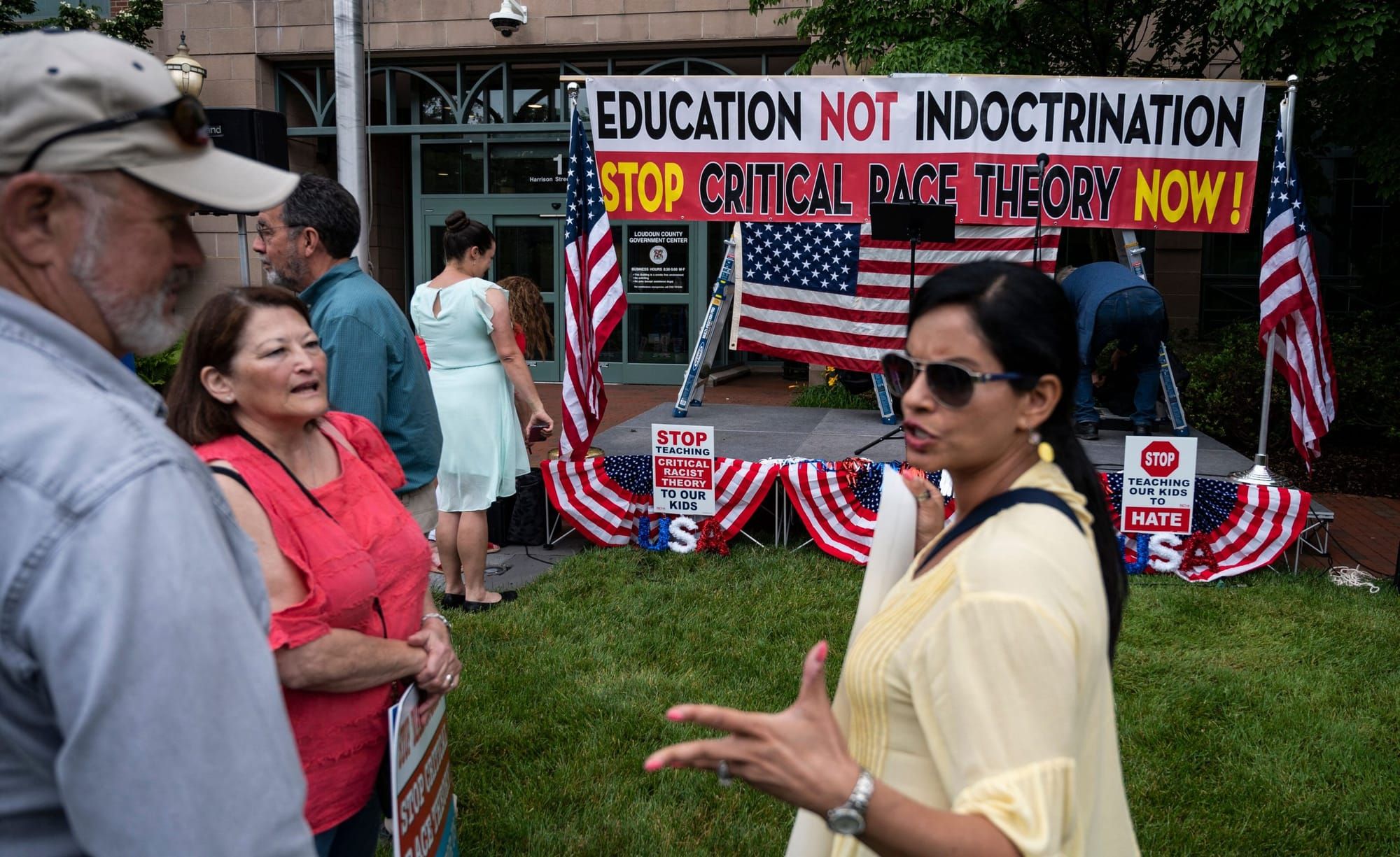 a woman with black hair tied back in a ponytail wearing a white flowing shirt stands in front of a sign that says "Education