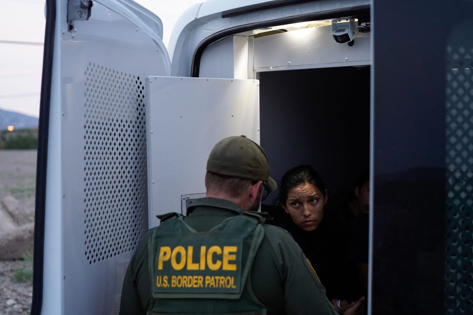 a woman with dark hair sits in the back of a van, whose doors are open. a border patrol agent faces her with their back to th