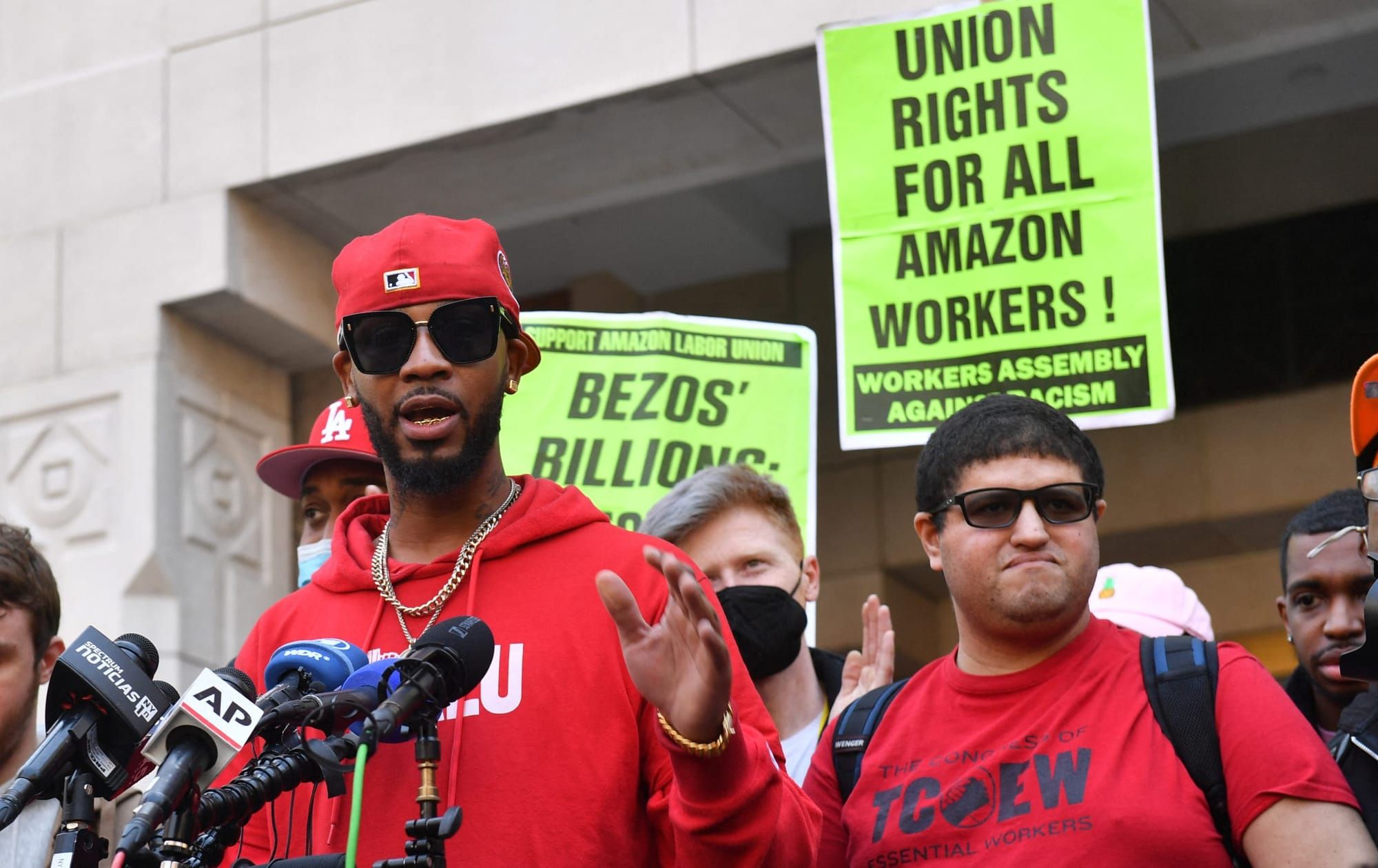 a black man in a red hoodie and baseball cap worn backwards stands at a microphone outside. behind him is a sign in green tha
