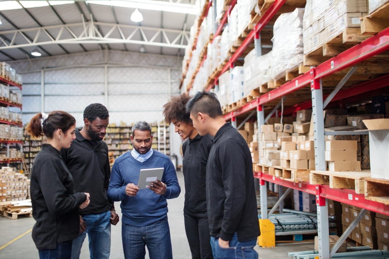 middle-aged man holds an iPad as four younger colleagues stand around him in a warehouse