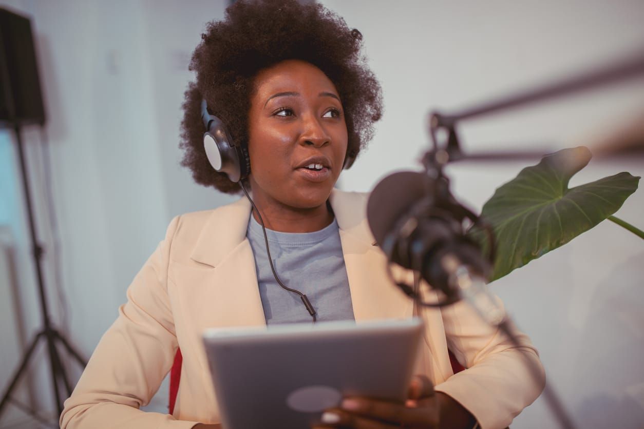 a young Black woman sits at a desk behind a podcast microphone while reading off a notepad
