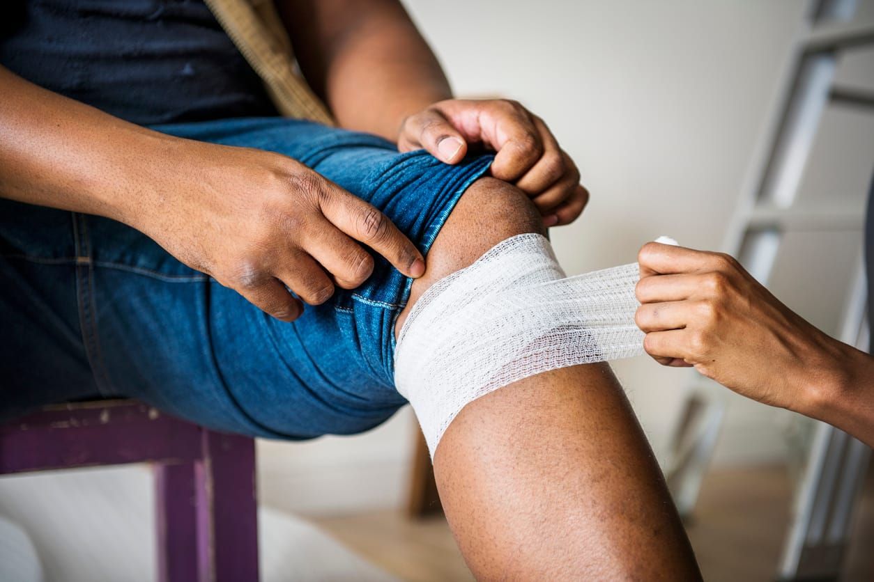 a Black person points below their knee as another Black person wraps a bandage around their leg