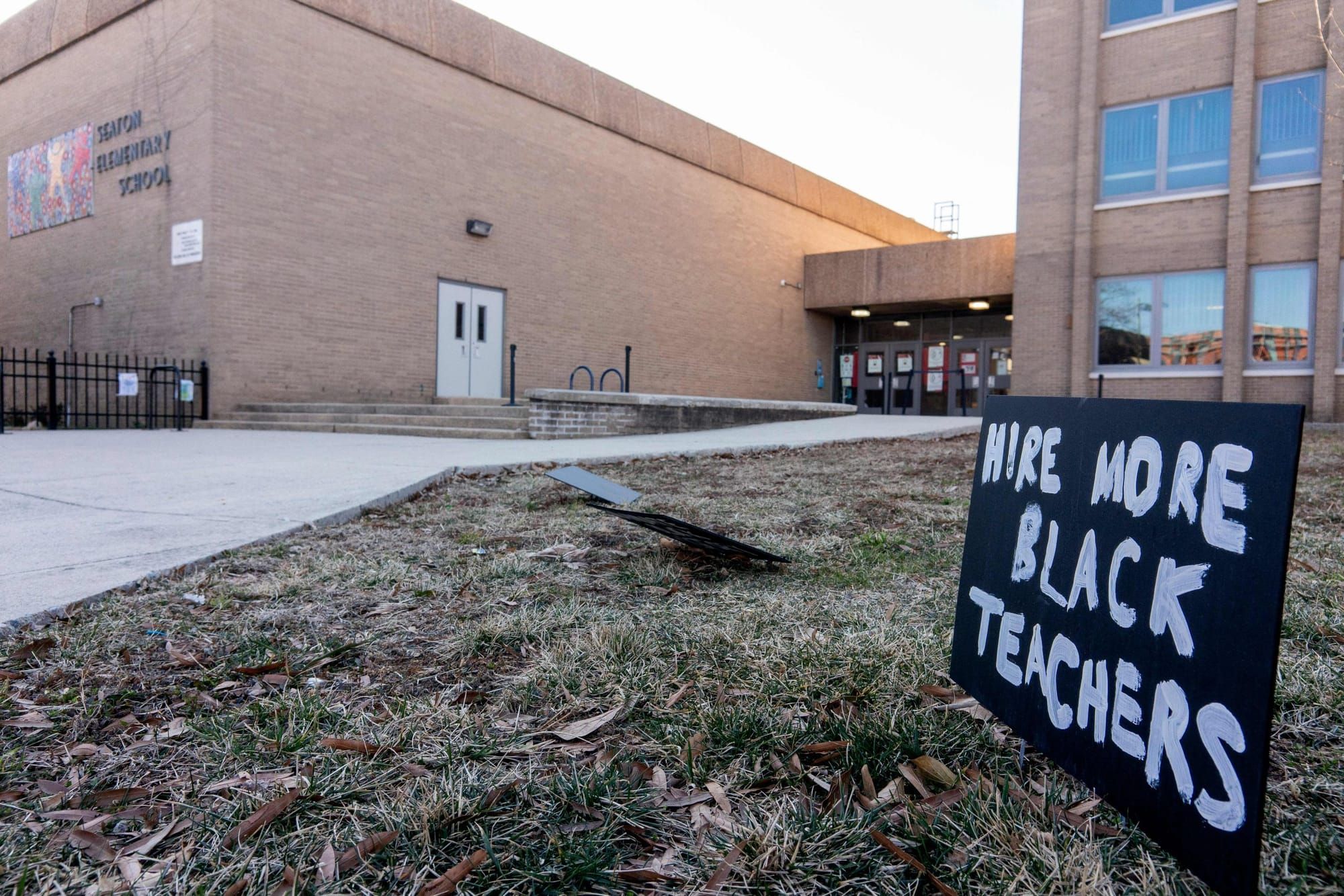 a sign reading "hire more Black teachers" stands outside a brick school building