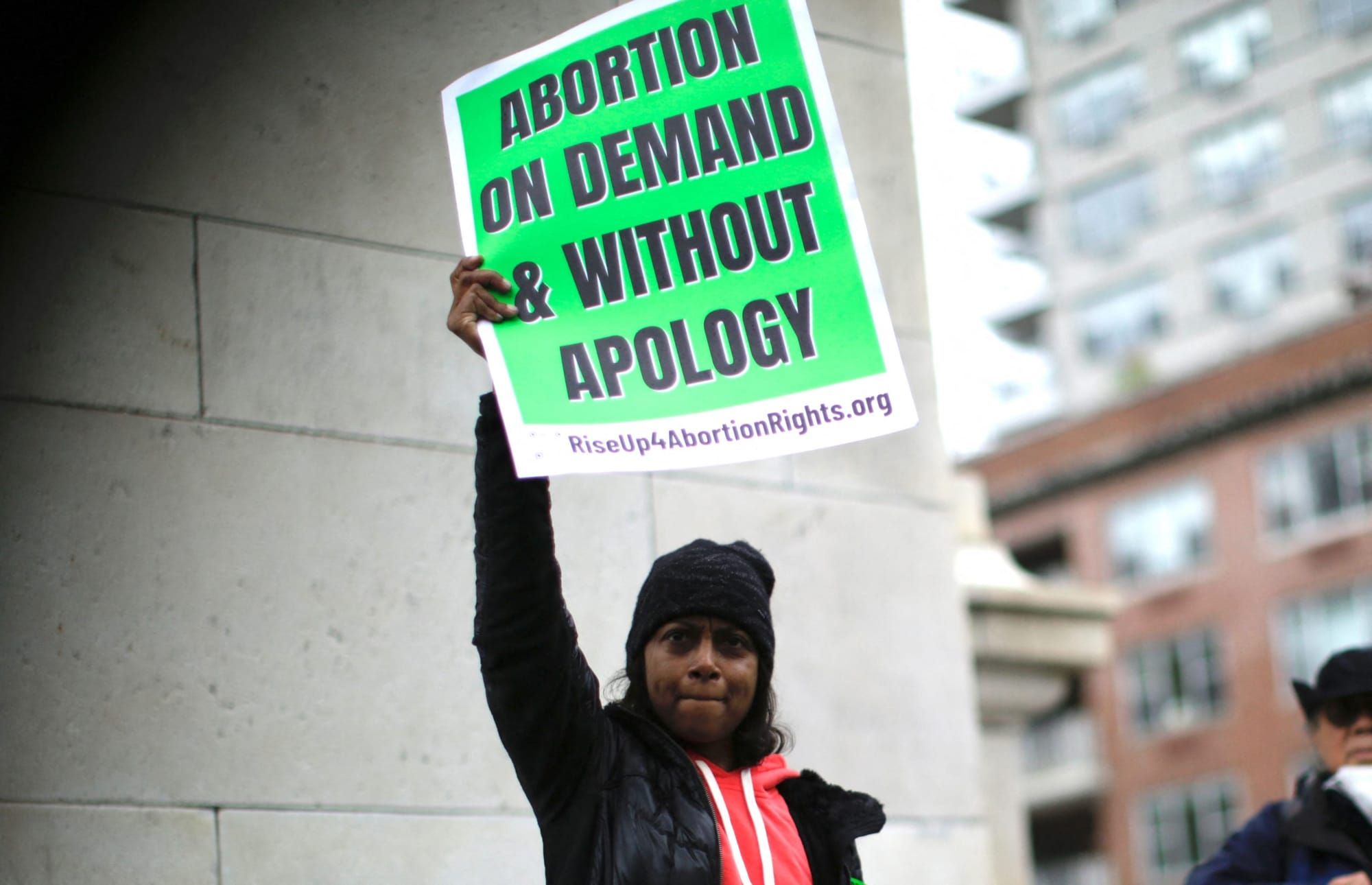 a Black femme person wearing black clothing while holding a green sign reading "abortion on demand and without apology"