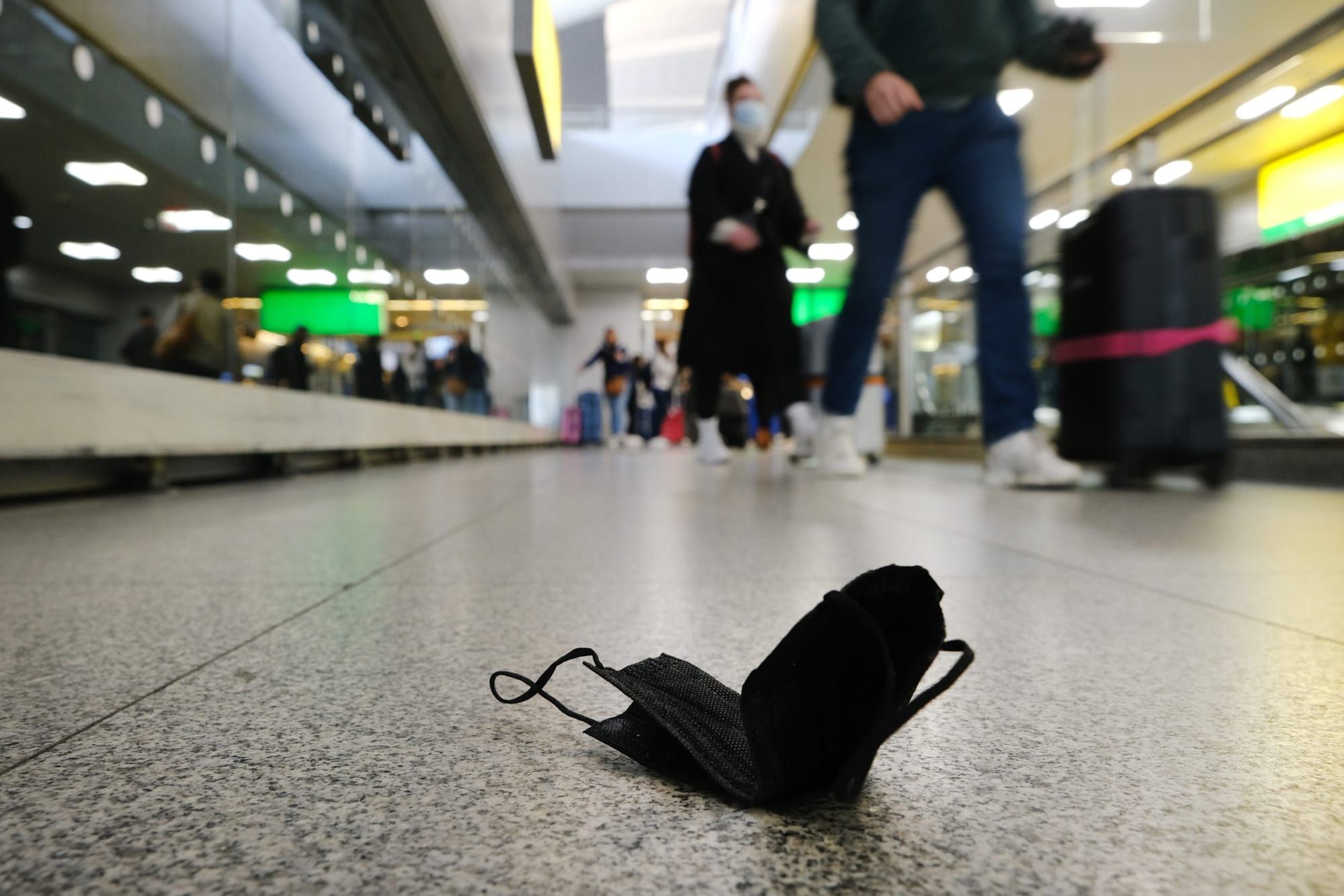 a black face mask lies abandoned on the ground in an airport terminal as people walk by