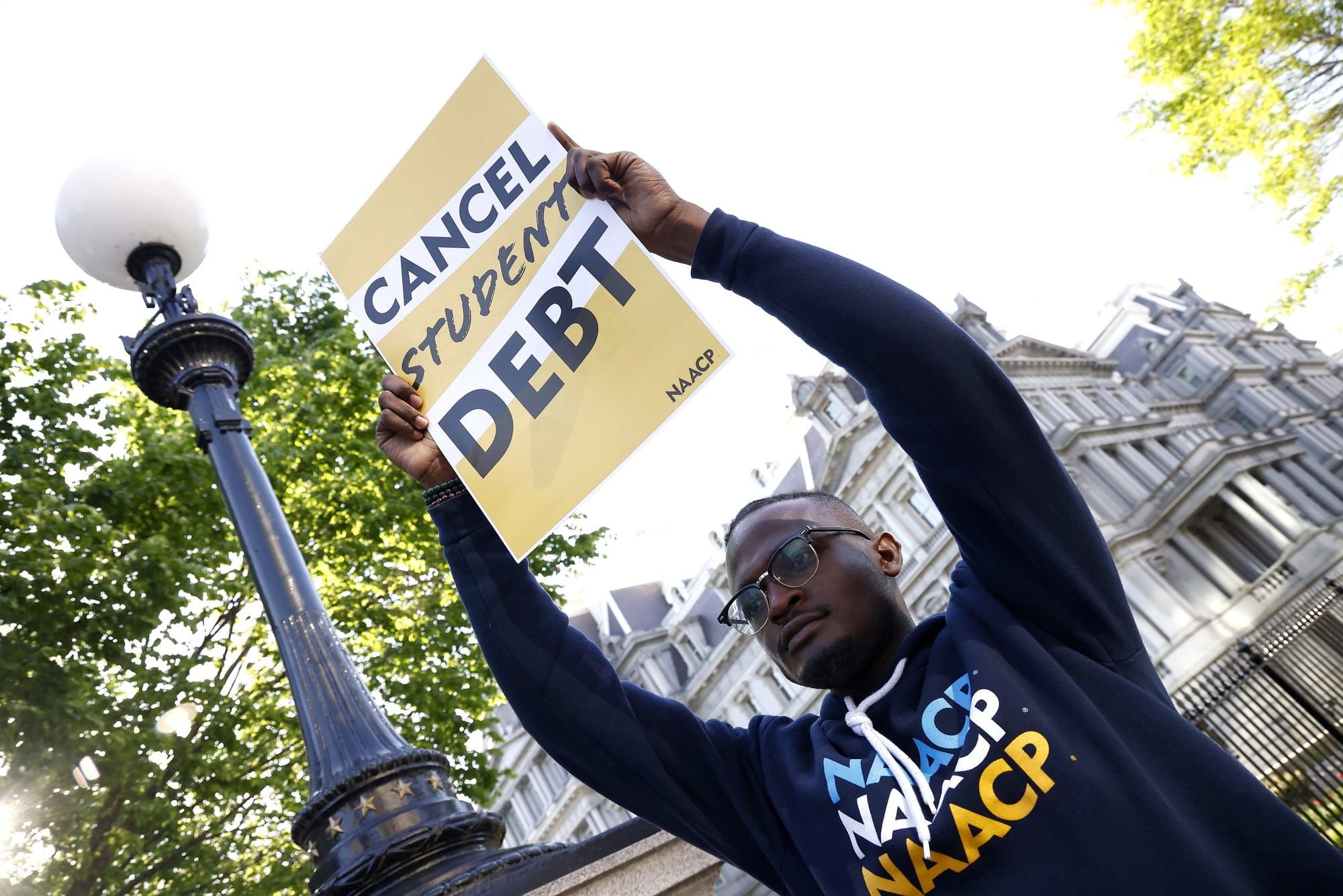 a Black person wearing an NAACP hoodie holds a sign above his head reading "cancel student debt"