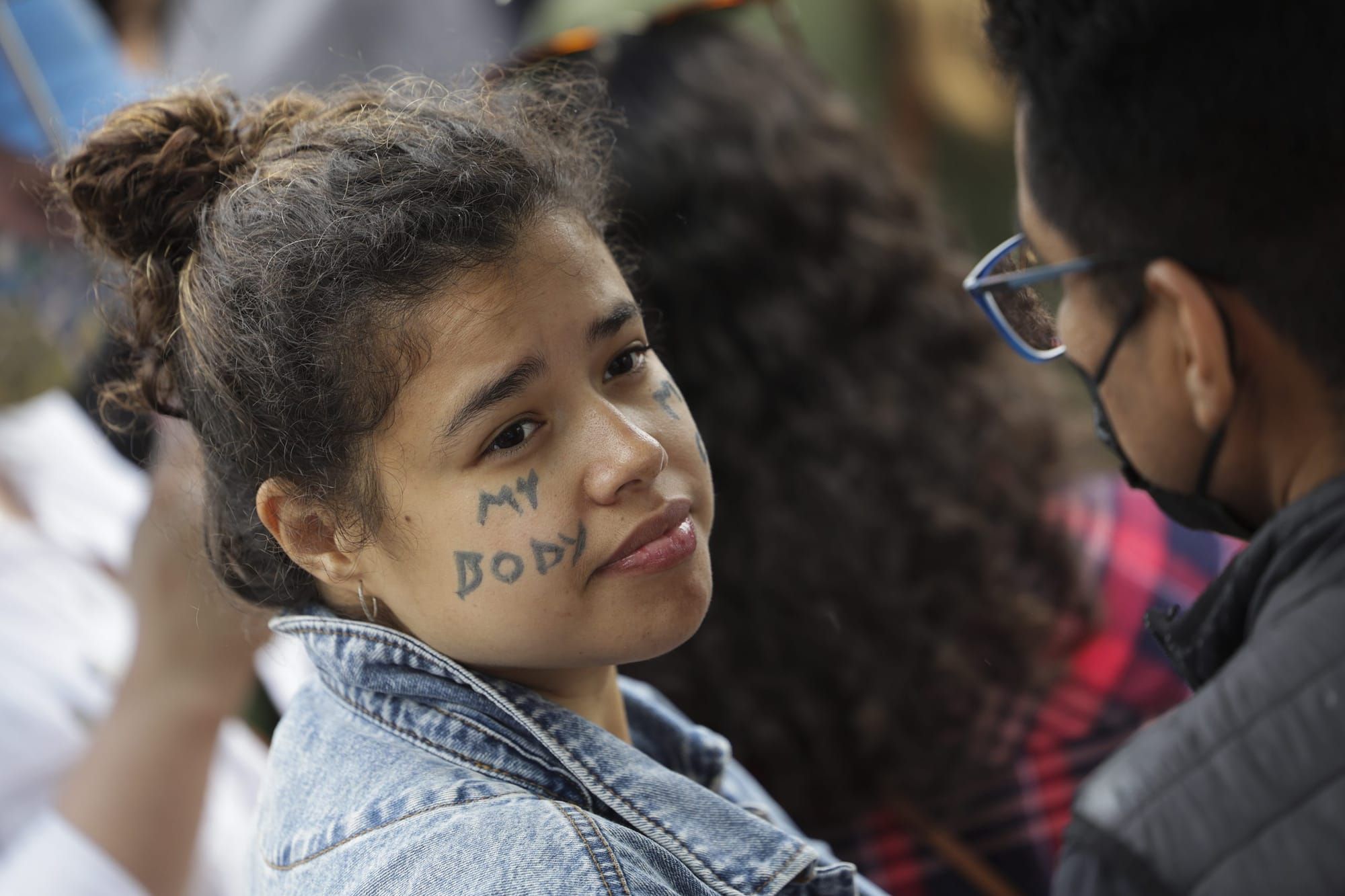 an adolescent girl looks over her shoulder toward the camera during a pro-abortion rally. "my body" is written on her cheek